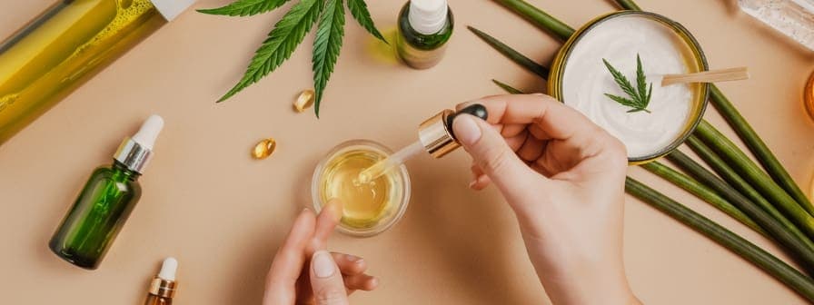 Overhead view of a table with different CBD products laid out, including cream, oil, and capsules, with a pair of hands in the middle using a dropper to apply CBD oil to hands for skin care benefits